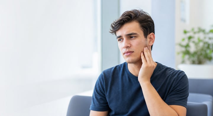 Young adult gently holding their jaw near the ear due to wisdom tooth pain in a bright, modern dental office.